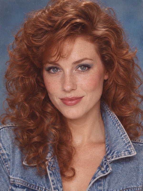 The same red-haired woman with freckles in the same pose with big curls, blue makeup and a denim jacket in an 80s portrait