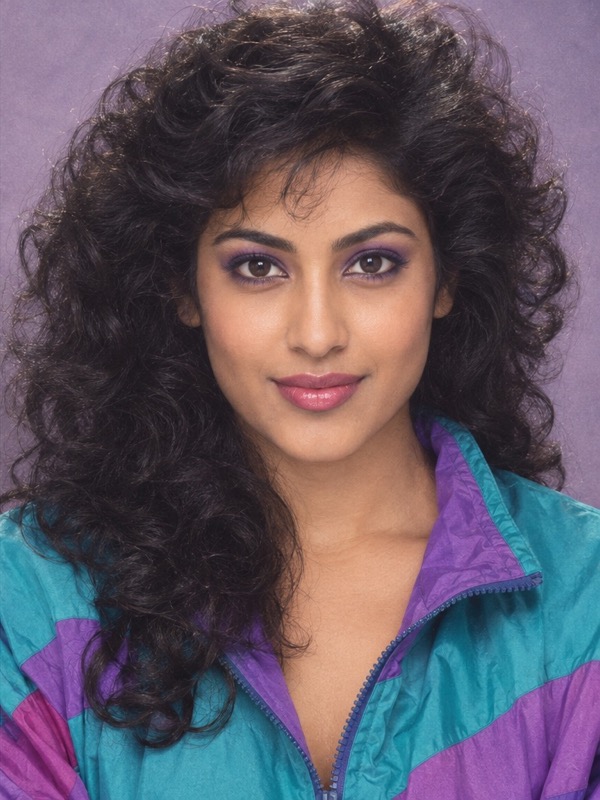 The same South Asian woman in the same pose with full curls, purple makeup and a colorful jacket in an 80s portrait